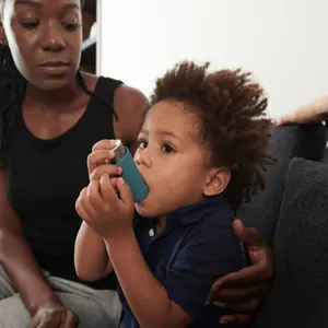 Worried mother looking at son using inhaler when having asthma attack at home