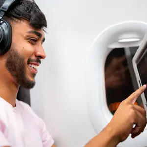 Latin American man in plane cabin using smart device listening to music on headphones.