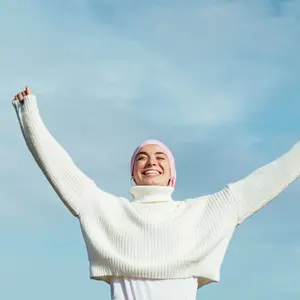 Happy young woman with cancer with arms up. She is wearing a pink scarf on her head and white jersey.