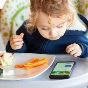 Toddler eats in the high chair while watching movies on the mobile phone.
