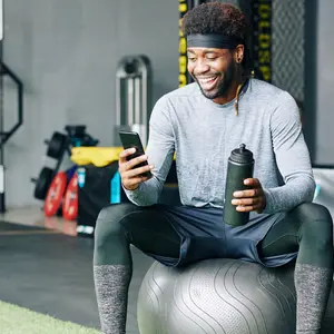 Excited young Black man sitting on fitness ball with bottle of water and checking social media on smartphone