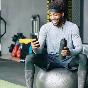 Excited young Black man sitting on fitness ball with bottle of water and checking social media on smartphone Excited young Black man sitting on fitness ball with bottle of water and checking social media on smartphone