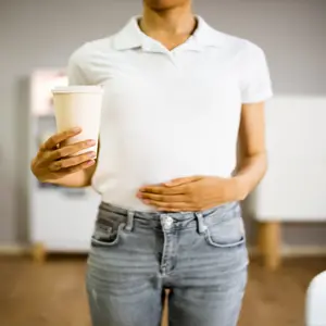 Person holding a cup of coffee with one hand and their stomach with another hand